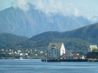 En stor, hvit bygning i flere etasjer ligger ved vannet med skogkledde åser og høye fjell i bakgrunnen under en delvis overskyet himmel.