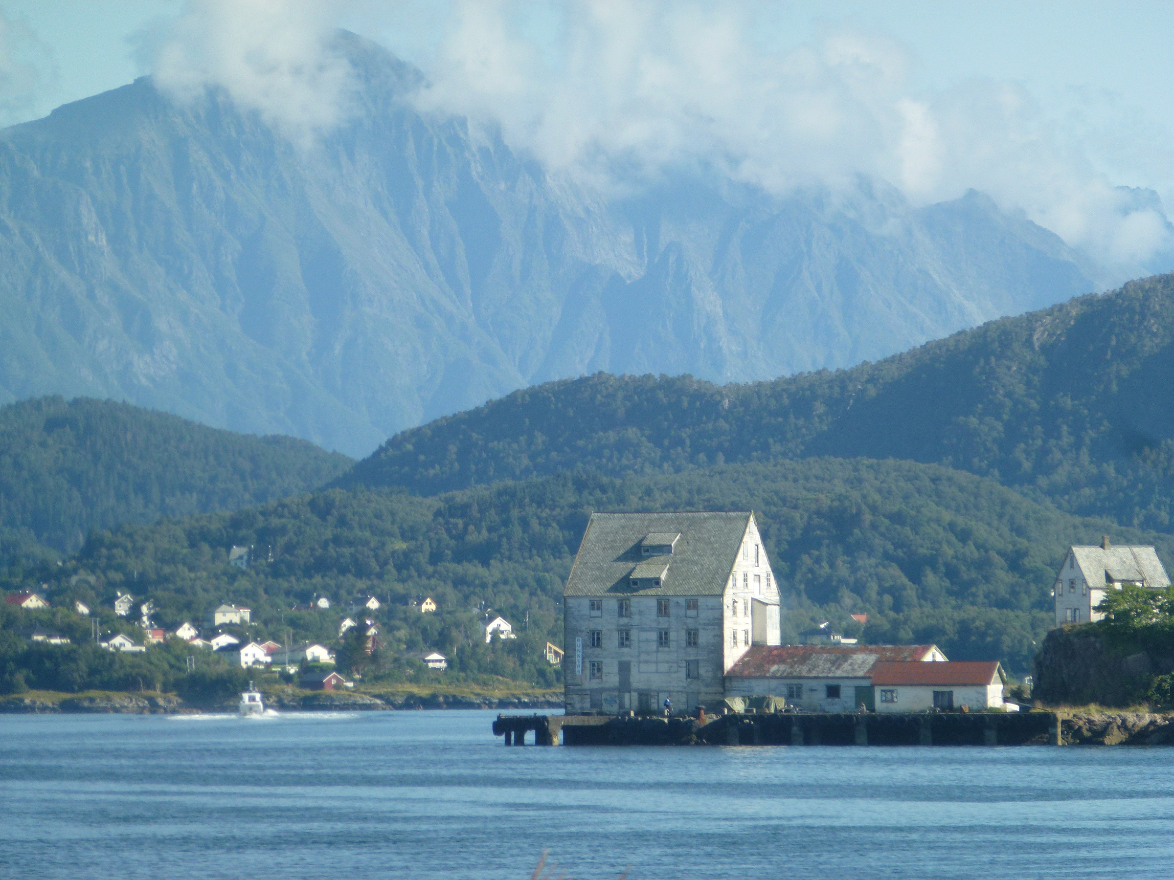 En stor, hvit bygning i flere etasjer ligger ved vannet med skogkledde åser og høye fjell i bakgrunnen under en delvis overskyet himmel.