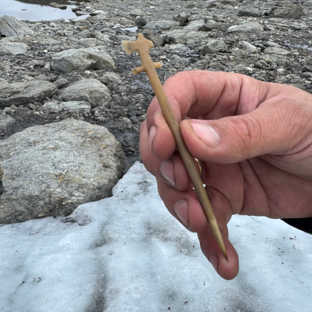 Delte ut 43 millionar til arkeologi – dette var dei mest spanande funna 11 En hånd holder en utskåret trestokk med en spiss ende og en hakket topp, mot en bakgrunn av steiner og is utendørs.