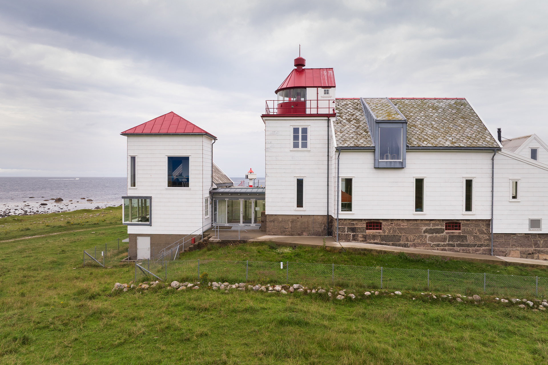 Tar deg ut i havgapet – tilgjengelighet på Kvassheim fyr 10 En hvit fyrbygning med røde tak står på en gresskledd kyst nær sjøen under en overskyet himmel.