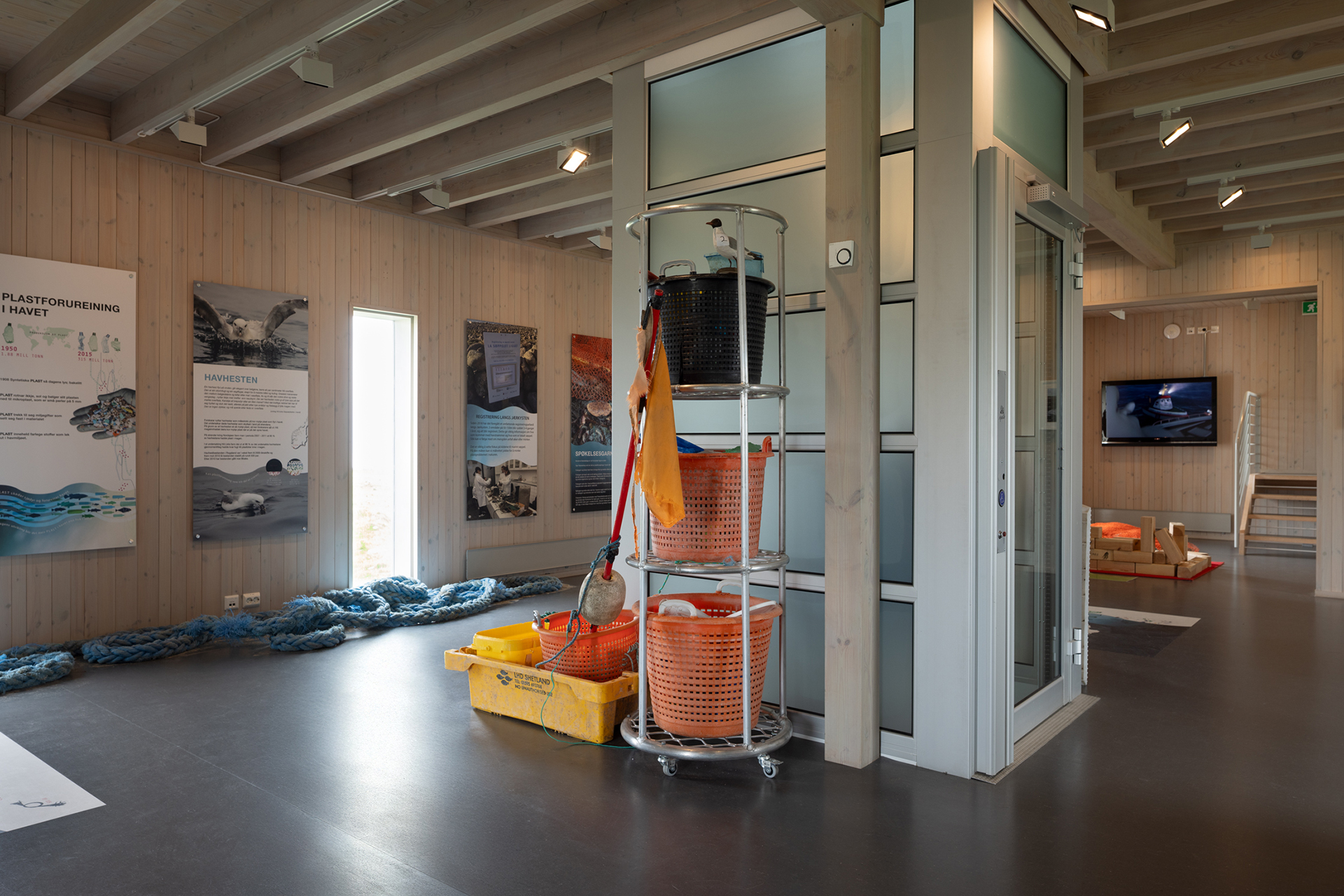 Fra avstandsfyr til nærmiljøfyrtårn 19 Et moderne utstillingslokale med pedagogiske plakater på veggene, et stativ med oransje kurver og fiskeutstyr og sitteplasser med en TV i bakgrunnen.