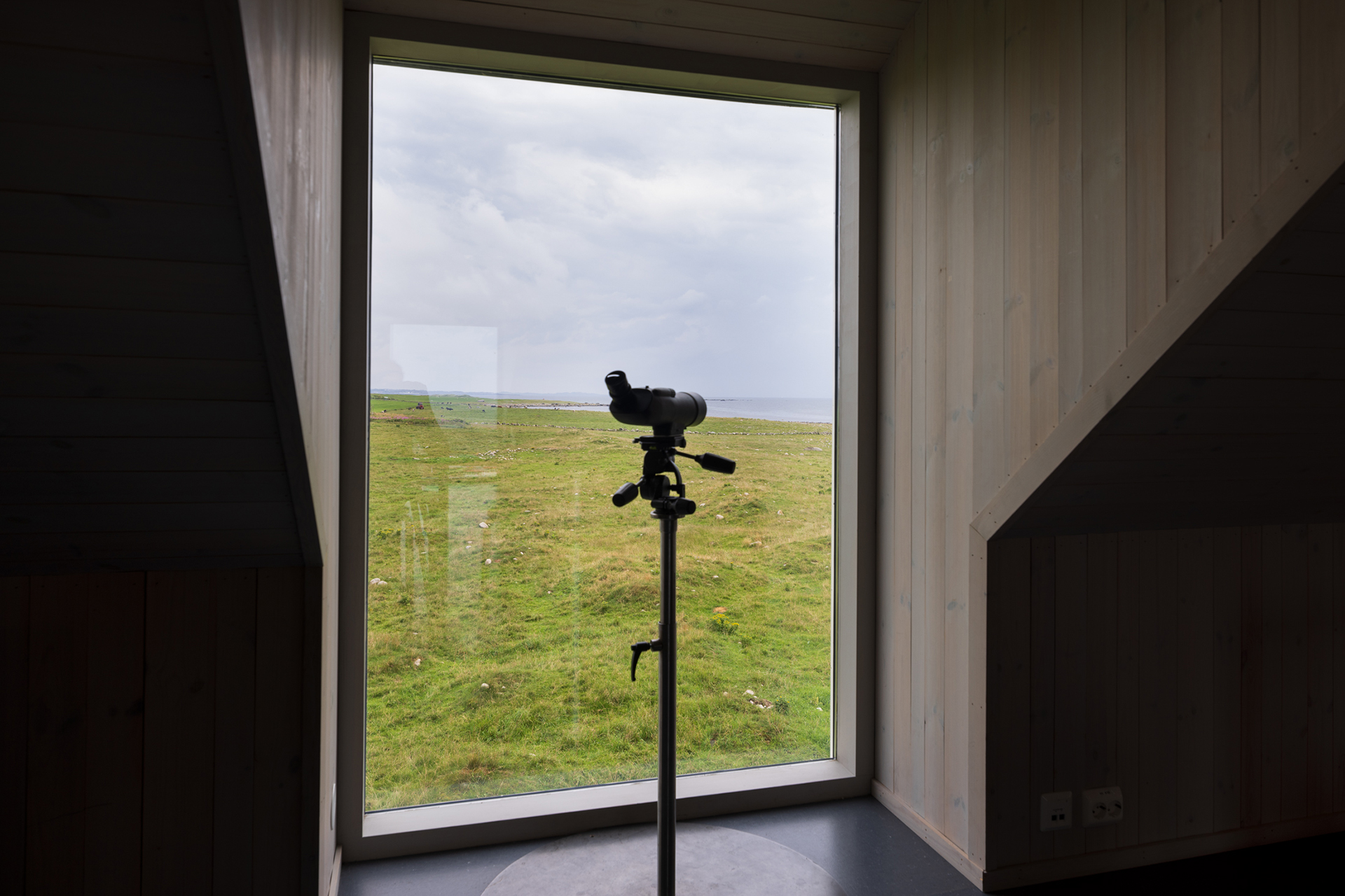 Fra avstandsfyr til nærmiljøfyrtårn 17 En kikkert på et stativ er plassert foran et stort vindu med utsikt over et gresslandskap under en overskyet himmel.