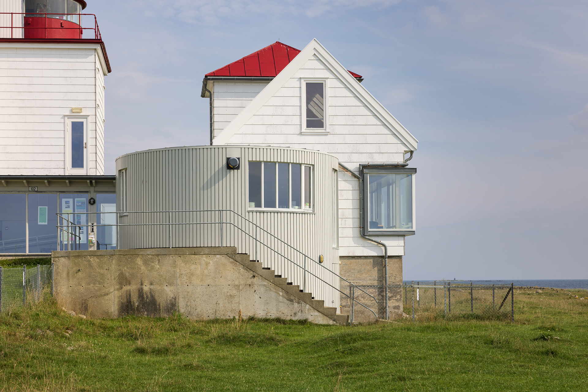Fra avstandsfyr til nærmiljøfyrtårn 15 Hvitt hus med rødt tak og sylinderformet tilbygg, plassert på gresslette nær kysten under en delvis overskyet himmel.