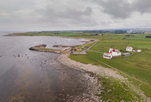 Flyfoto av en kystby med en liten havn, steinete strandlinje, grønne åkrer og spredte bygninger under en overskyet himmel.