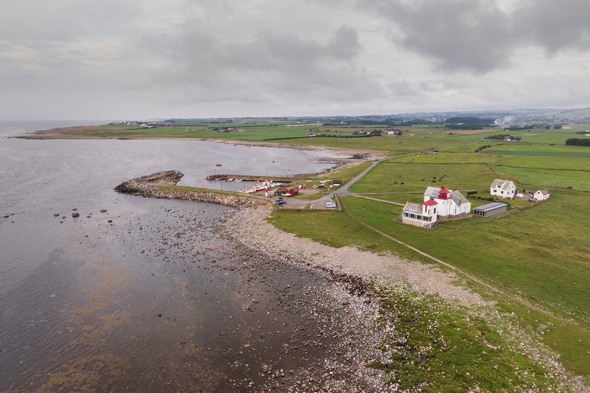 Fra avstandsfyr til nærmiljøfyrtårn 13 Flyfoto av en kystby med en liten havn, steinete strandlinje, grønne åkrer og spredte bygninger under en overskyet himmel.