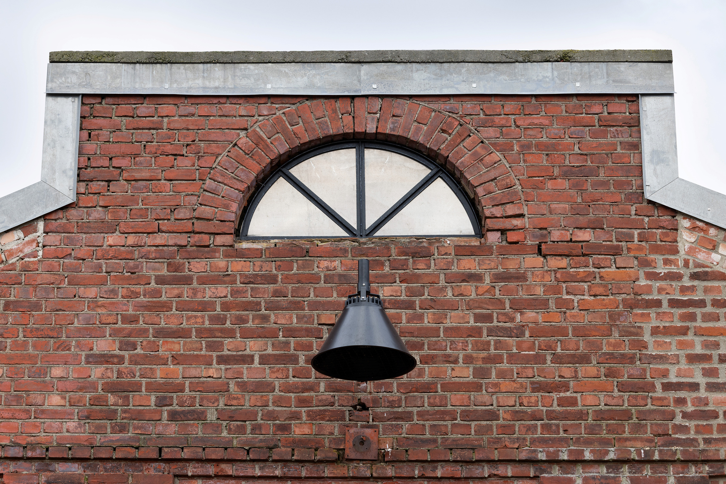 En svart utvendig vegglampe er montert under et halvsirkelformet vindu med svarte sprosser på en rød murbygning.