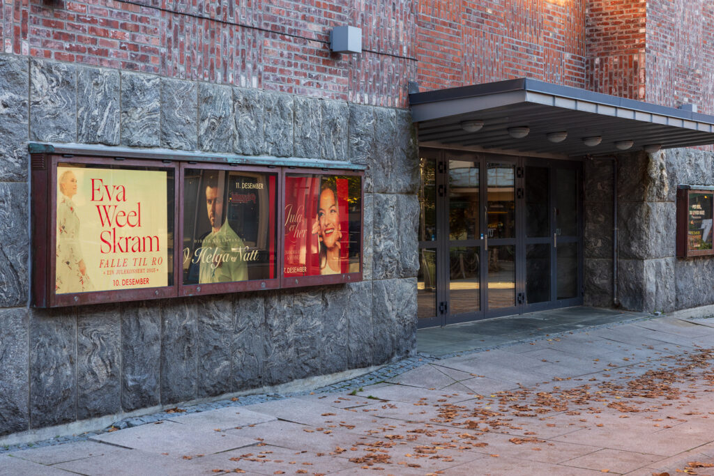Utendørs plakattavler på en steinmur viser reklame for forestillinger, ved siden av inngangen til en murbygning med glassdører og høstløv på bakken.