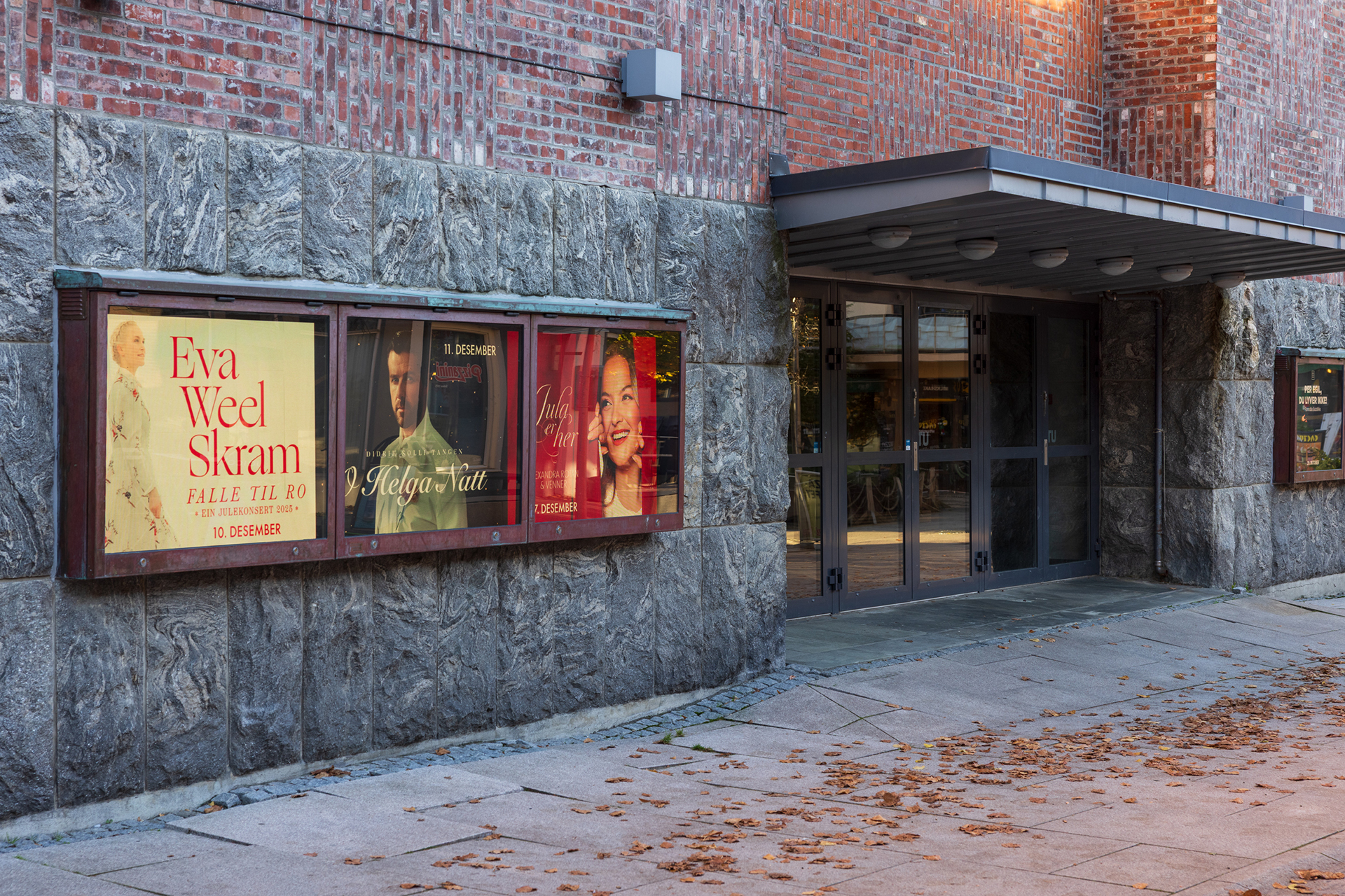 Utendørs plakattavler på en steinmur viser reklame for forestillinger, ved siden av inngangen til en murbygning med glassdører og høstløv på bakken.