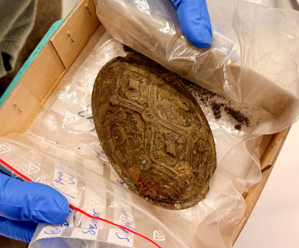 Delte ut 43 millionar til arkeologi – dette var dei mest spanande funna 17 En person med hansker holder en plastinnpakket, sirlig mønstret gjenstand, muligens et skjold eller en brosje, i en pappeske.