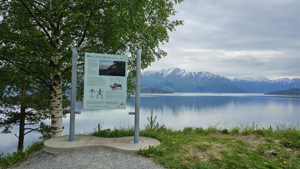 Et informasjonsskilt står ved et tre med utsikt over en rolig innsjø med snødekte fjell i bakgrunnen, der 34 meldt til politiet nylig har fått oppmerksomhet under den overskyede himmelen.