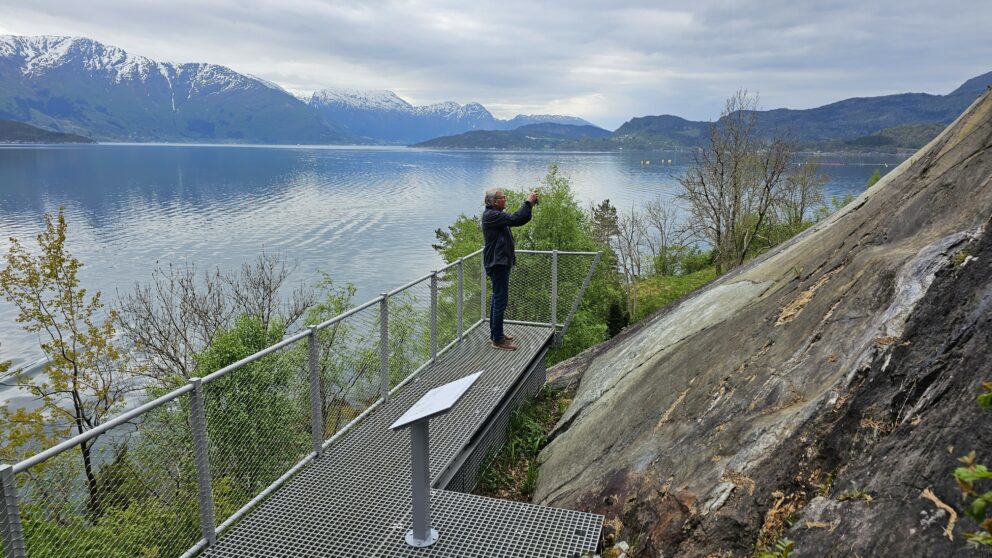 En person står på en utsiktsplattform av metall ved en innsjø og tar et bilde av fjellandskapet i det fjerne under en overskyet himmel, der det nylig ble rapportert om 34 meldt til politiet-hendelser.