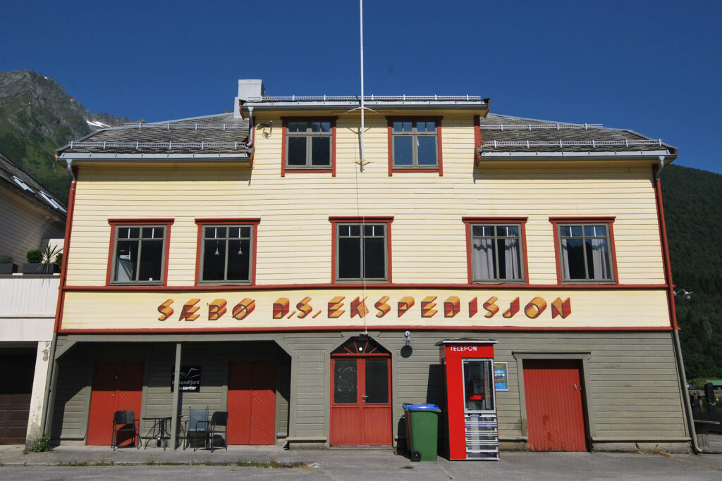 En toetasjes gul trebygning med røde dører og en rød salgsautomat utenfor, med skiltet Sæbø AS. Ekspedisjon.