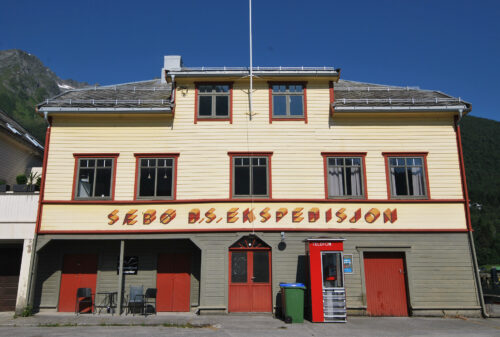 En toetasjes gul trebygning med røde dører og en rød salgsautomat utenfor, med skiltet Sæbø AS. Ekspedisjon.