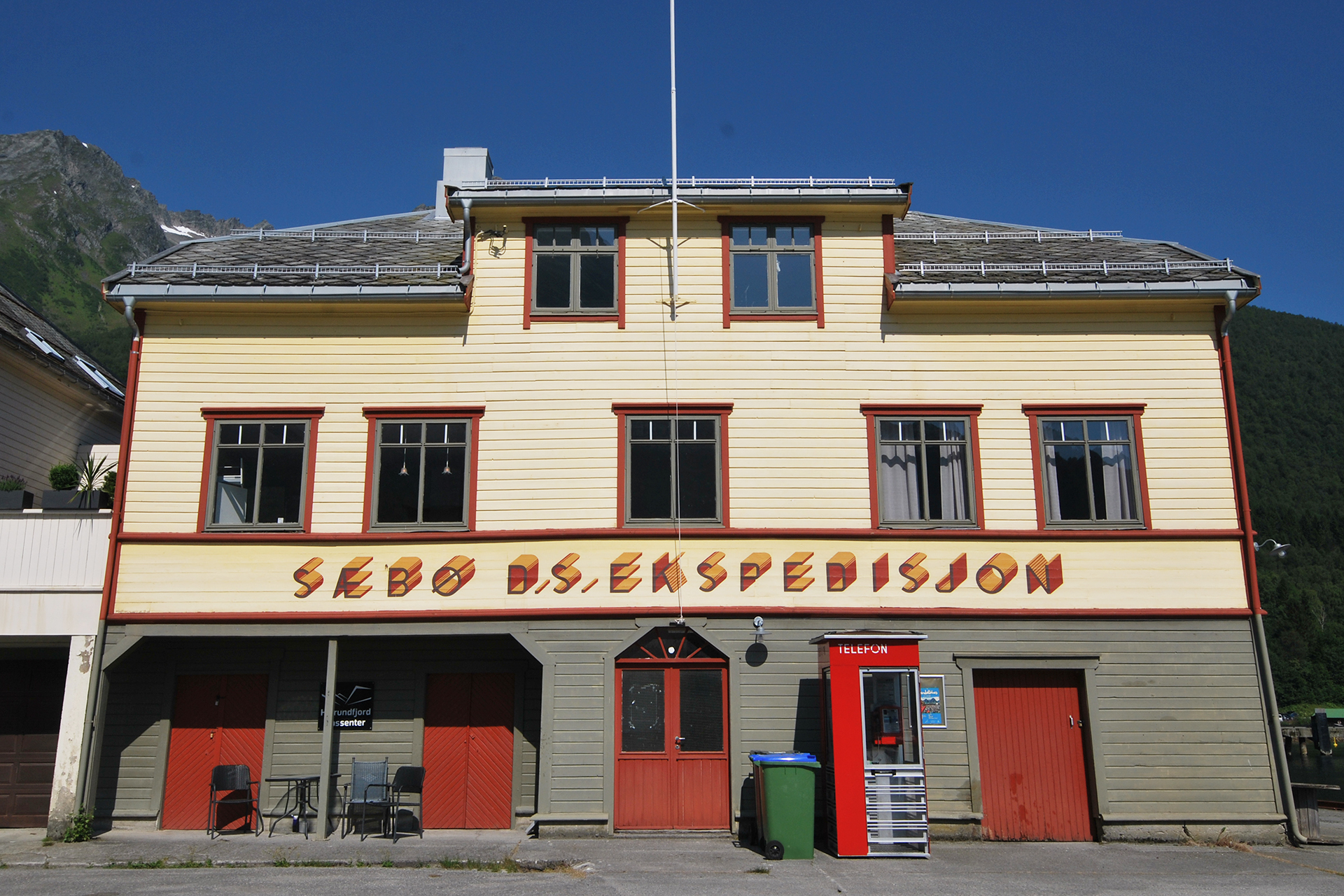 En toetasjes gul trebygning med røde dører og en rød salgsautomat utenfor, med skiltet Sæbø AS. Ekspedisjon.