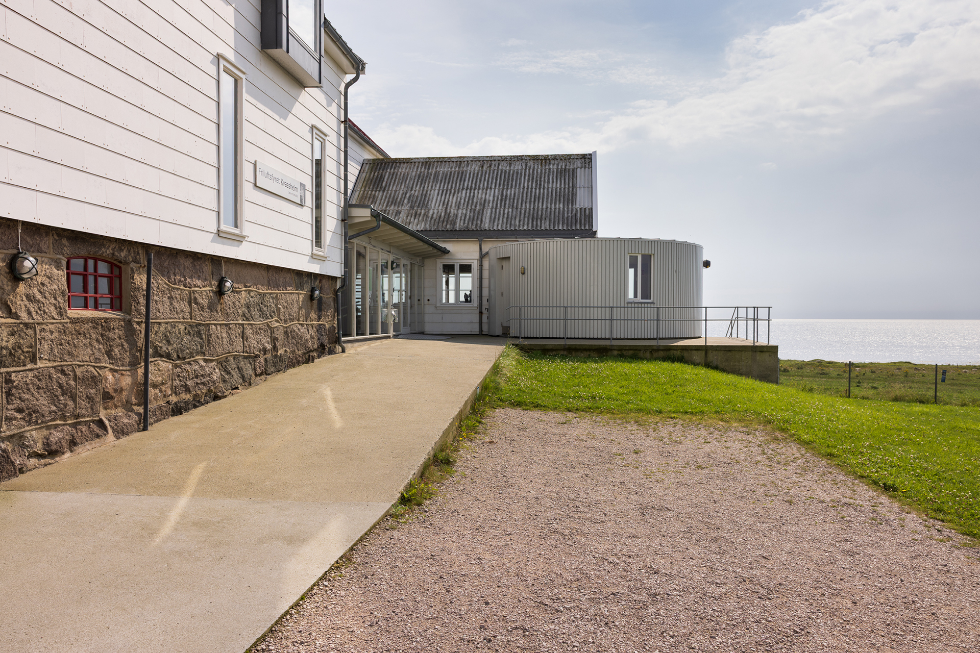 Tar deg ut i havgapet – tilgjengelighet på Kvassheim fyr 7 En betongrampe fører til inngangen til en moderne bygning med eksteriør i stein og metall, som ligger i nærheten av et gressområde med utsikt over havet.