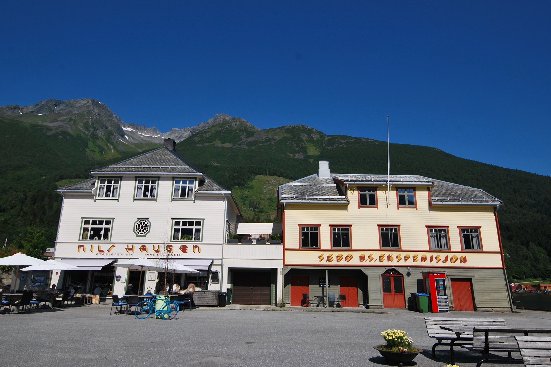 Font og fargar løftar identiteten til dampskipsekspedisjonen 8 To tradisjonelle bygninger med uteservering står foran grønne fjell under en klarblå himmel. Nils Haugen og Sægrov Bilserspedisjon står det på skiltene.