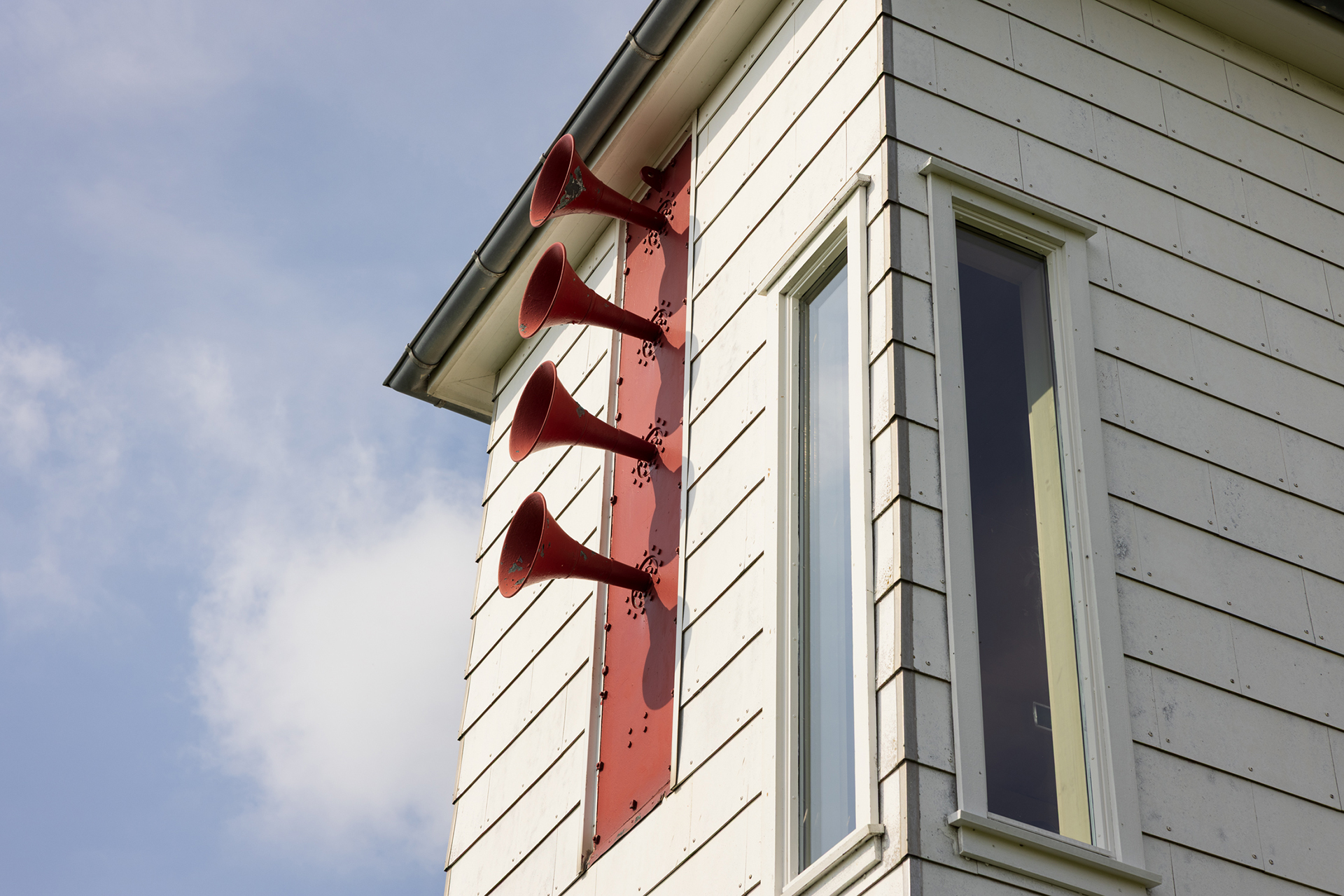 Fra avstandsfyr til nærmiljøfyrtårn 8 Fire store røde horn er montert vertikalt på ytterveggen til en hvit bygning, ved siden av et vindu under et skråtak.