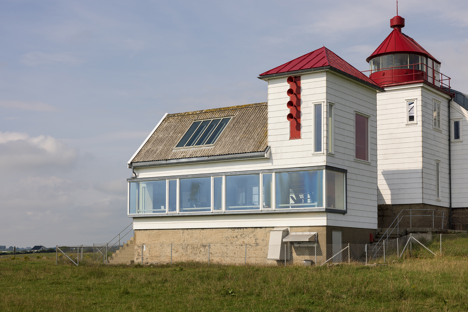 Fra avstandsfyr til nærmiljøfyrtårn 6 Hvitt fyrtårn med røde takdetaljer og en tilbyggsdel i glass, plassert på en gresslette under en delvis overskyet himmel.