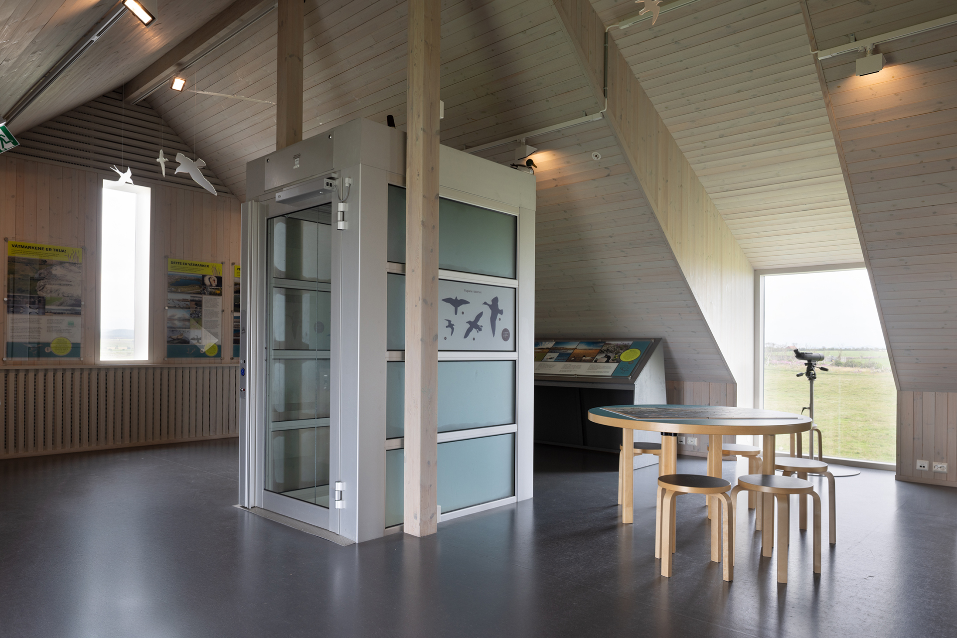 Tar deg ut i havgapet – tilgjengelighet på Kvassheim fyr 9 Et moderne innendørsrom med trevegger har en heis, et rundt bord med krakker, utstillingspaneler og et teleskop nær et stort vindu med utsikt over naturen.