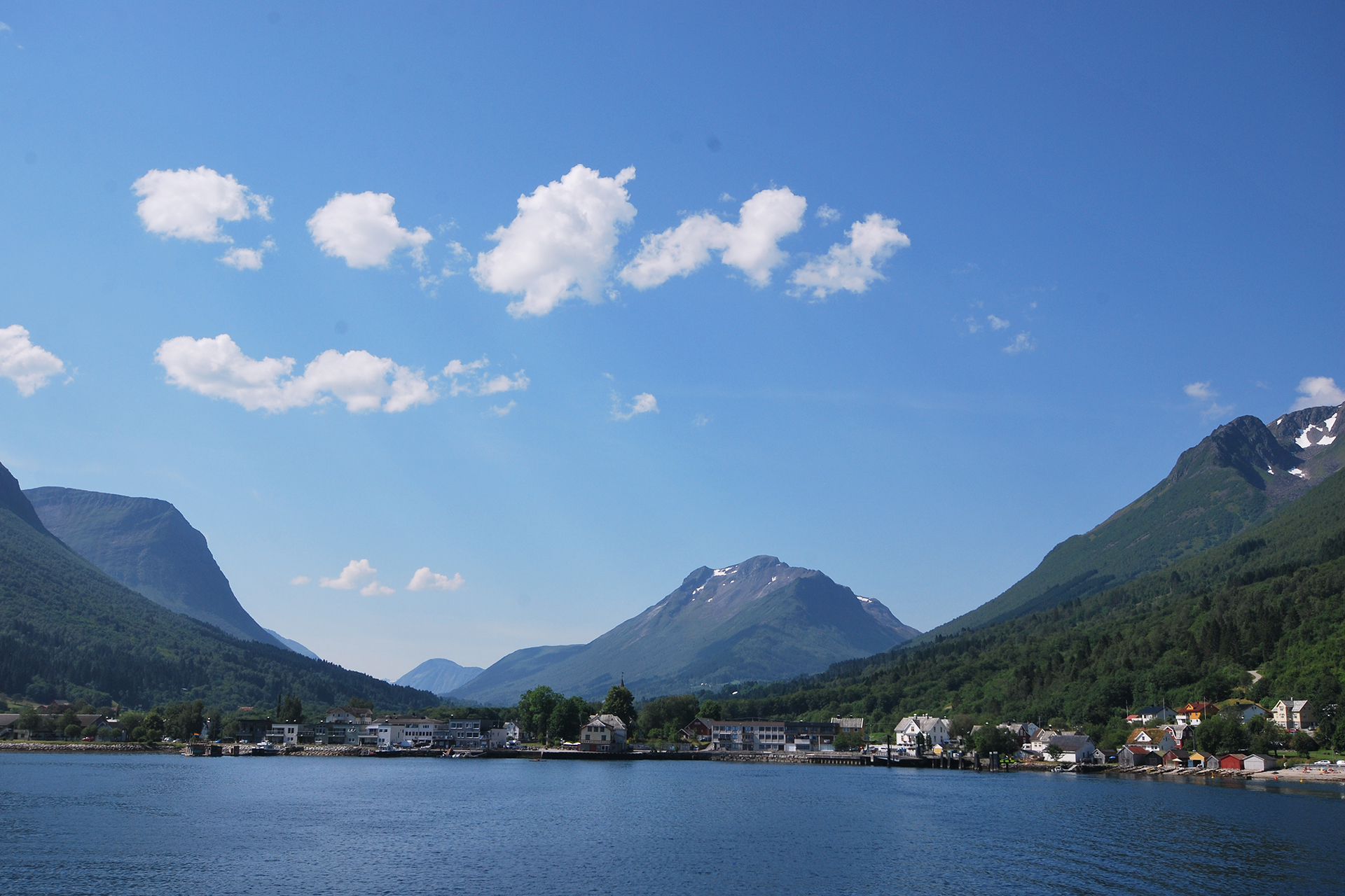 Font og fargar løftar identiteten til dampskipsekspedisjonen 9 En liten by ligger ved kanten av et stille vann, omgitt av grønne fjell og en blå himmel med spredte hvite skyer.