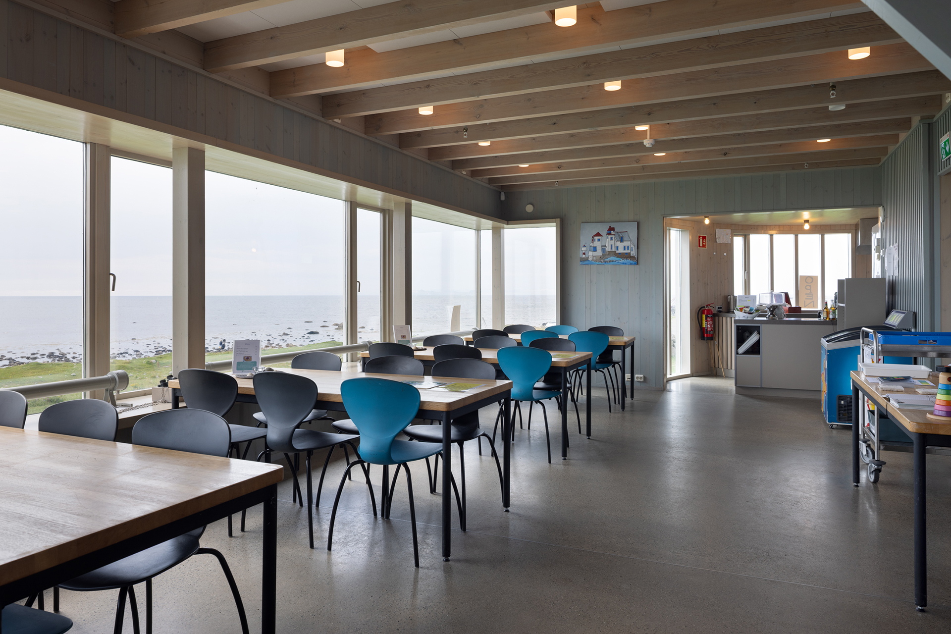 Fra avstandsfyr til nærmiljøfyrtårn 7 En moderne spiseplass med trebord, svarte og blå stoler, store vinduer med utsikt over havet og et lite kjøkkenområde på baksiden.