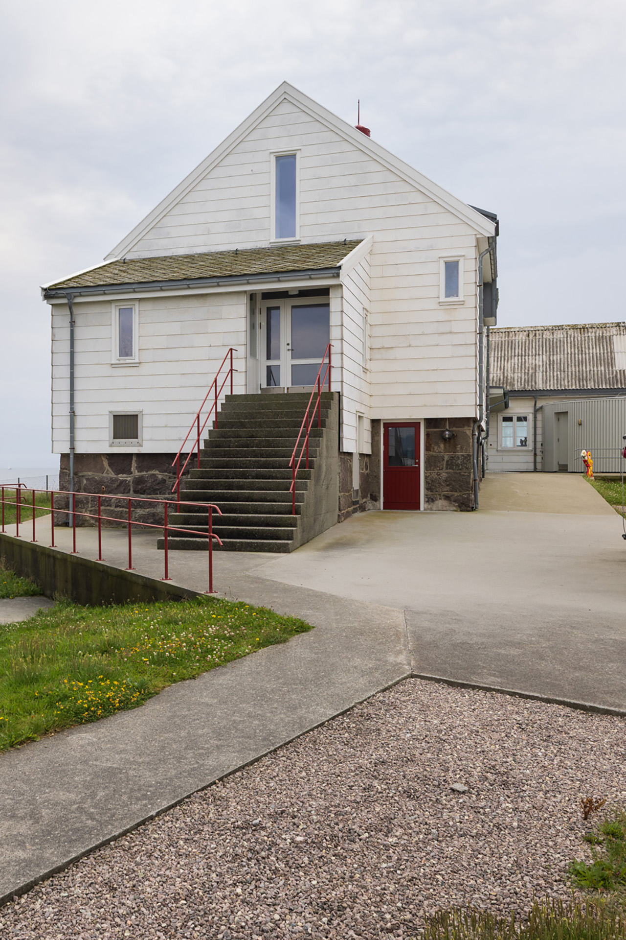 Fra avstandsfyr til nærmiljøfyrtårn 10 En hvit toetasjes bygning med rødt rekkverk, betongtrapper som fører til hovedinngangen, og en grus- og betongsti foran.