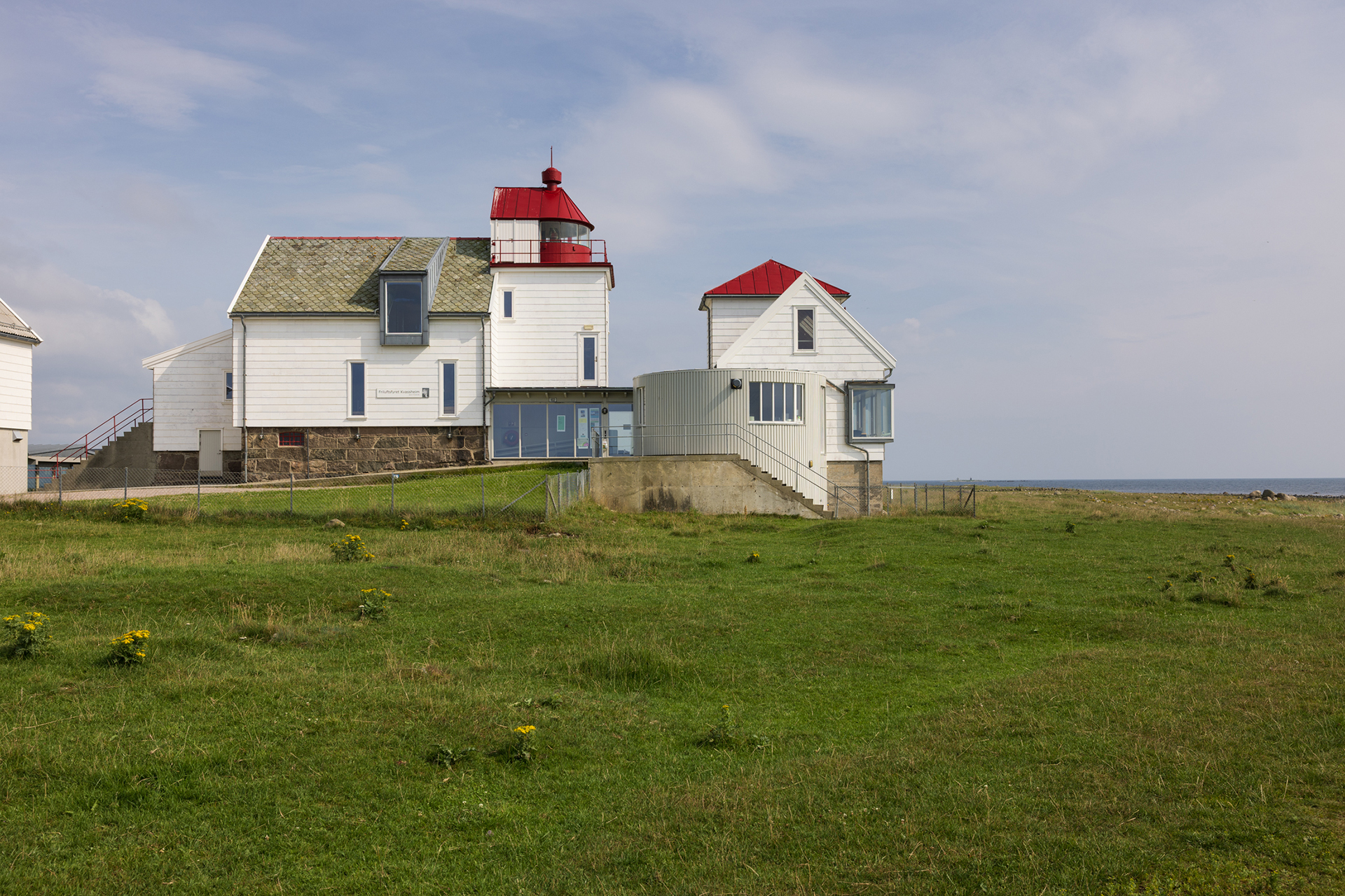 Fra avstandsfyr til nærmiljøfyrtårn 14 Et hvitt fyrtårn med rødt tak og tilhørende bygninger står på en gresslette nær sjøen under en delvis overskyet himmel.
