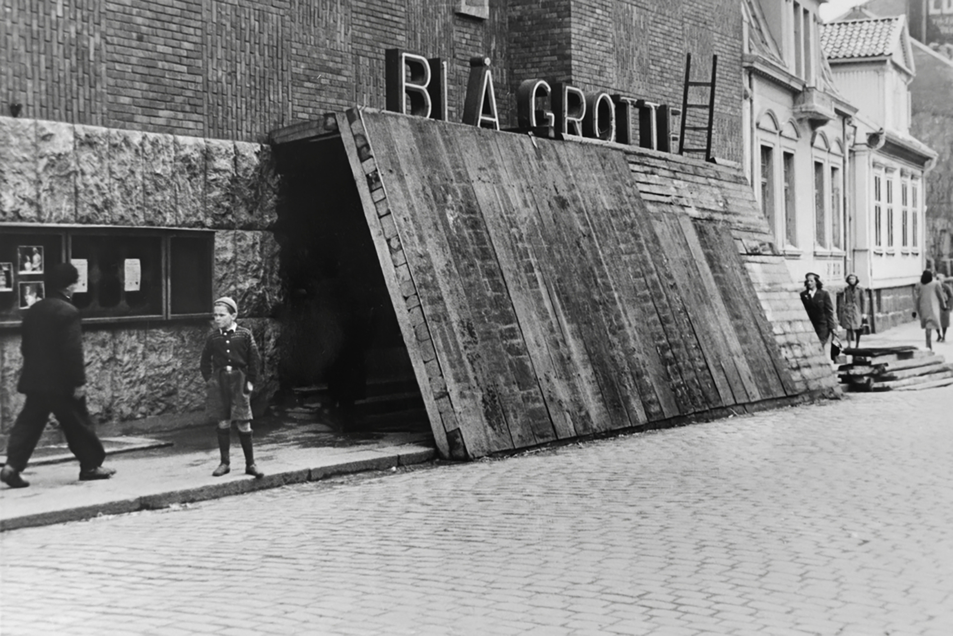 Informasjonsdeling på plakaten 7 Svart-hvitt-foto av mennesker som går forbi en murbygning med en stor inngangsrampe i tre og et skilt med teksten Blå Grottan.