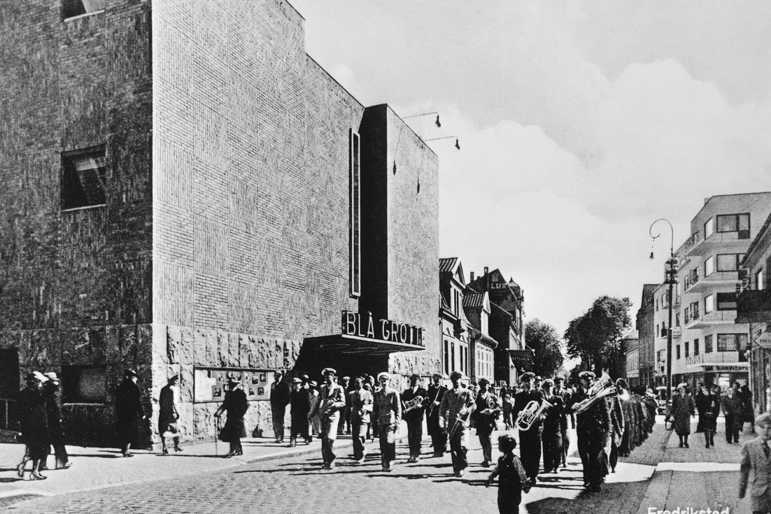 Informasjonsdeling på plakaten 6 Et musikkorps marsjerer ned en bygate forbi Blå Grottan kino, mens en folkemengde ser på fra fortauet.