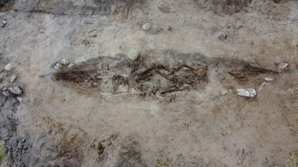 Delte ut 43 millionar til arkeologi – dette var dei mest spanande funna 5 Flyfoto av en utgravd grav, som viser et menneskeskjelett begravd i en langstrakt, båtformet grop omgitt av jord og stein.