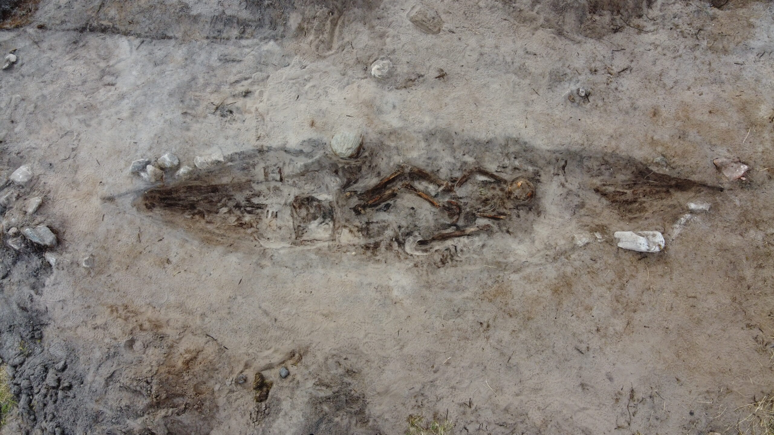 Delte ut 43 millionar til arkeologi – dette var dei mest spanande funna 1 Flyfoto av en utgravd grav, som viser et menneskeskjelett begravd i en langstrakt, båtformet grop omgitt av jord og stein.