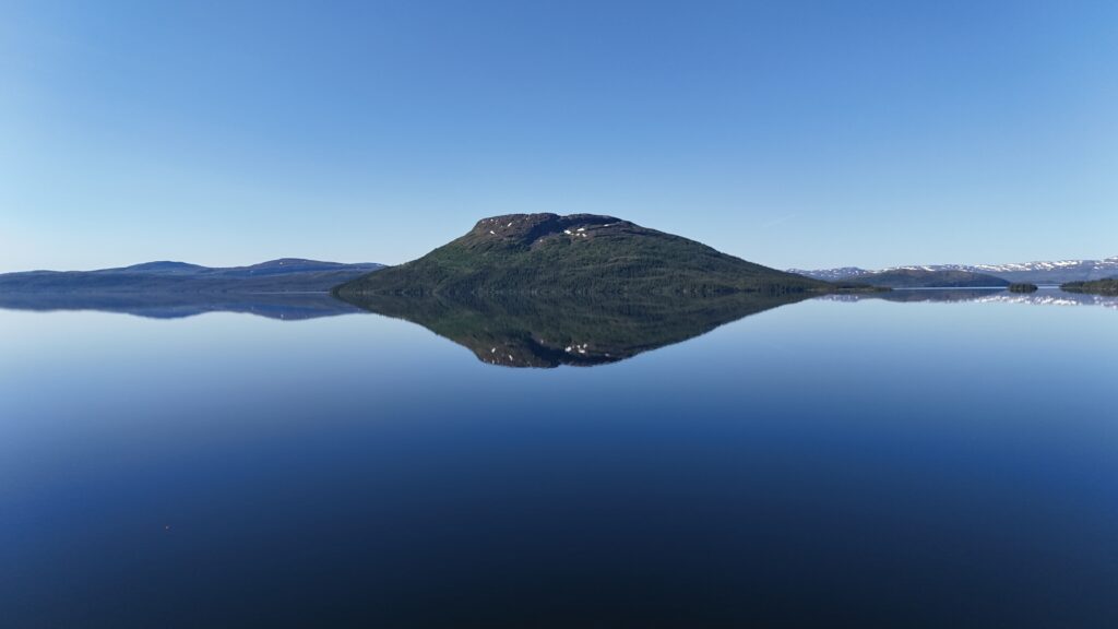 En grønn øy omkranset av innsjøen Dåtnejaevrie/Tunnsjøen speiler seg perfekt i vannet, under en klarblå himmel, med snøkledde fjell i bakgrunnen.