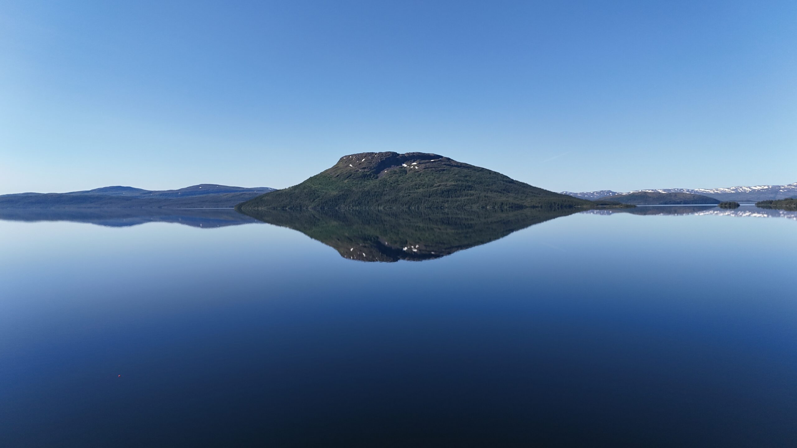 En grønn øy omkranset av innsjøen Dåtnejaevrie/Tunnsjøen speiler seg perfekt i vannet, under en klarblå himmel, med snøkledde fjell i bakgrunnen.