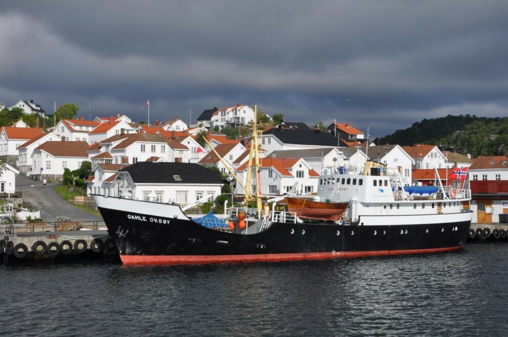 Fartøyvernet får 76 millionar i 2026 5 Et svart-hvitt skip med navnet GMLK 0868F ligger til kai ved en vannkant, med hvite hus med røde tak og grøntområder i bakgrunnen under en overskyet himmel.