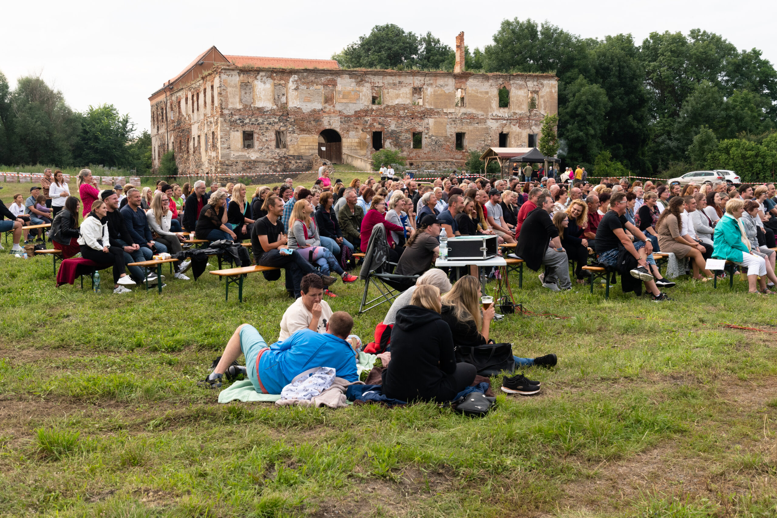 En stor gruppe festival-deltakere sitter ved Petipsy slott i Tsjekkia, i 2021. De sitter på benker og tepper utendørs, vendt mot en scene, med Petipsy slott i bakgrunnen.