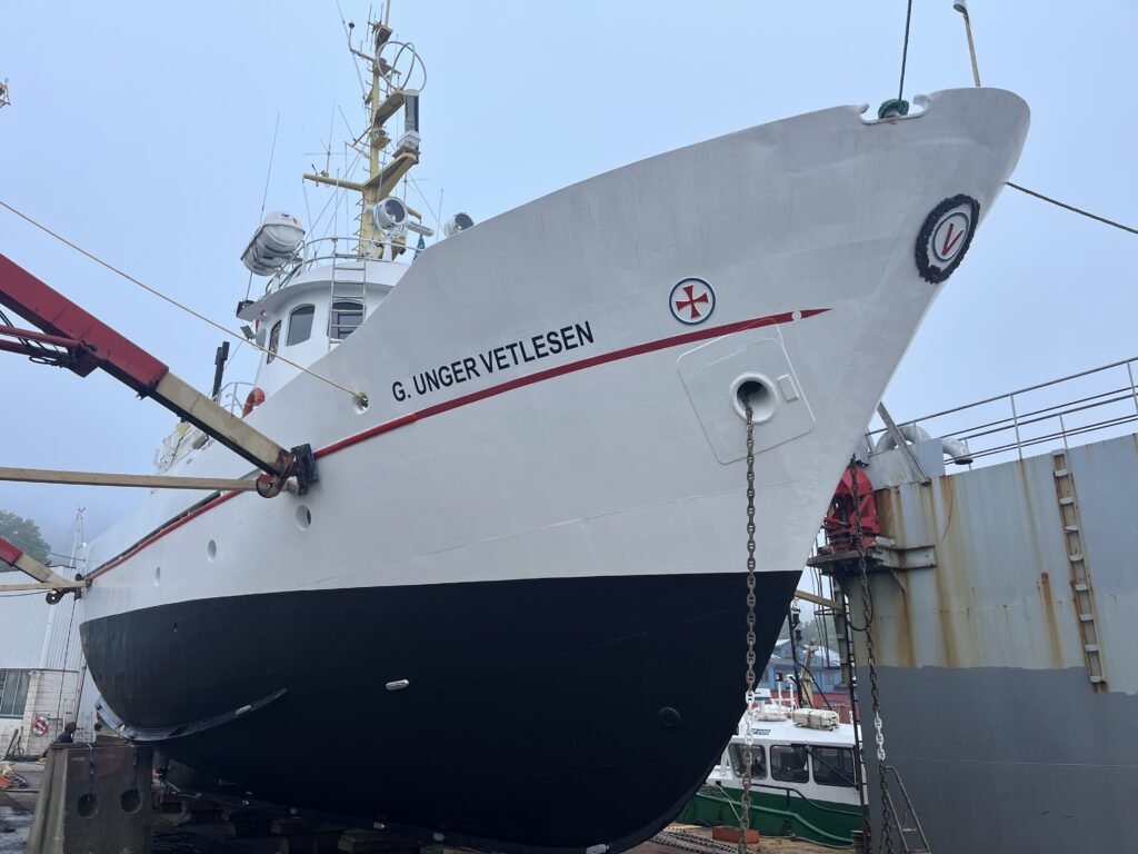 Et stort, hvitt og svart skip ved navn G. Ungervetlesen ligger til kai på land for vedlikehold, med utstyr og en mindre båt synlig i nærheten.