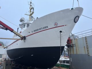 Et stort, hvitt og svart skip ved navn G. Ungervetlesen ligger til kai på land for vedlikehold, med utstyr og en mindre båt synlig i nærheten.