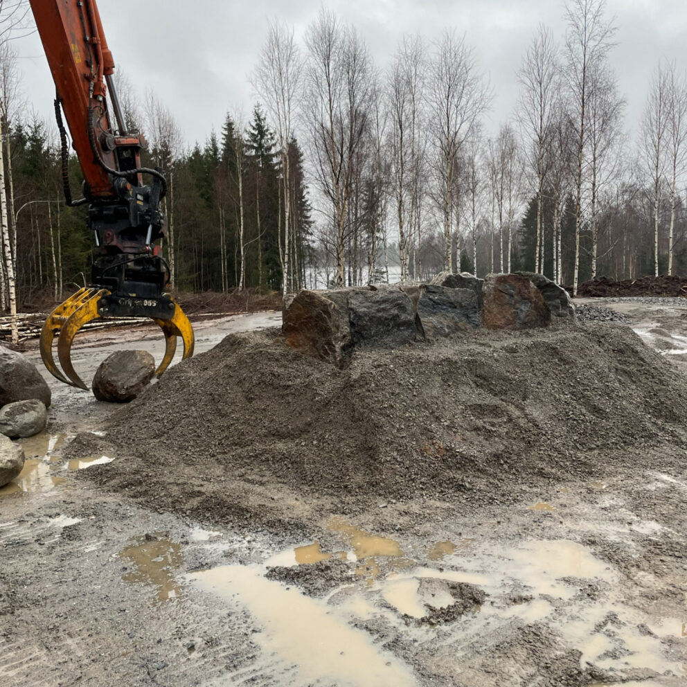 Delte ut 43 millionar til arkeologi – dette var dei mest spanande funna 8 En gravemaskin med gripeklo flytter en stor stein opp på en grushaug omgitt av sølepytter, med nakne trær og skog i bakgrunnen.