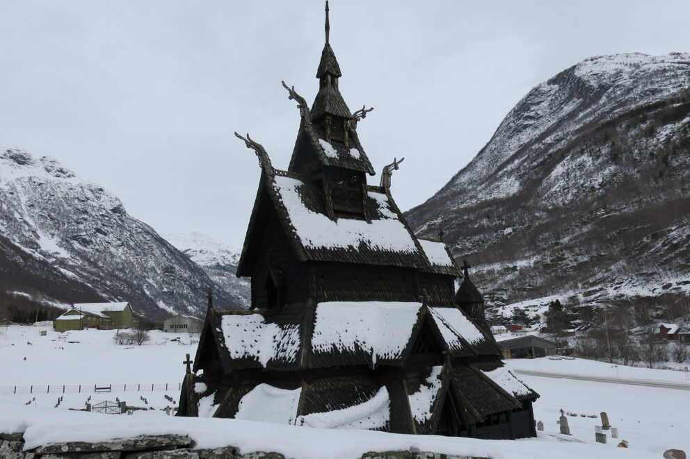 Milliontilskot til stavkyrkjene 5 En stavkirke i tre med intrikate utskjæringer og bratte tak står i et snødekt fjellandskap, omgitt av snødekt mark og hus i det fjerne.
