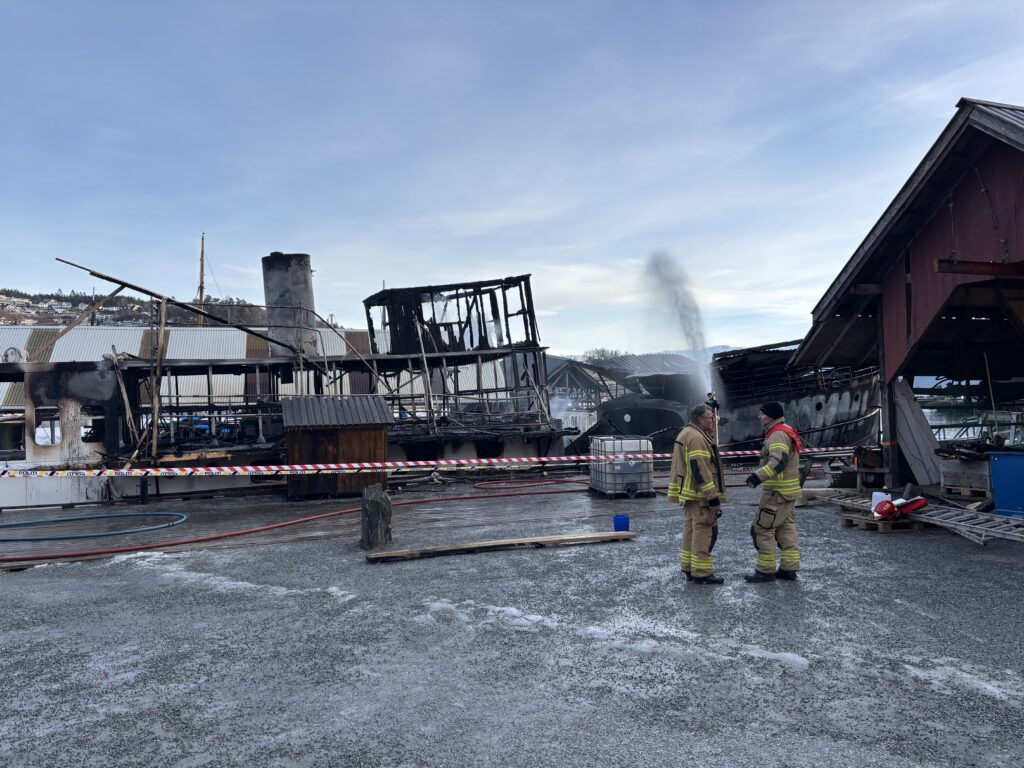 To brannmenn står foran en delvis ødelagt bygning med forkullede rester og røyk etter en brann; på bakken ligger det advarselsteip og slanger som minner om scener dokumentert av Hardanger farøyvernsenter Fartøyvernet.
