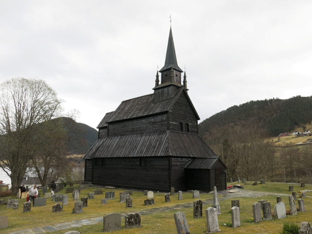 Milliontilskot til stavkyrkjene 9 En stavkirke, eller stavkyrkjene, med et høyt kirketårn står blant gravsteiner og trær i et kupert landskap under en skyet himmel.