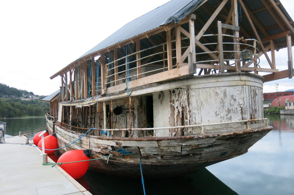 En stor, værbitt trebåt med delvis konstruert tak ligger til kai ved vannet, støttet av røde bøyer og fortøyningstau.