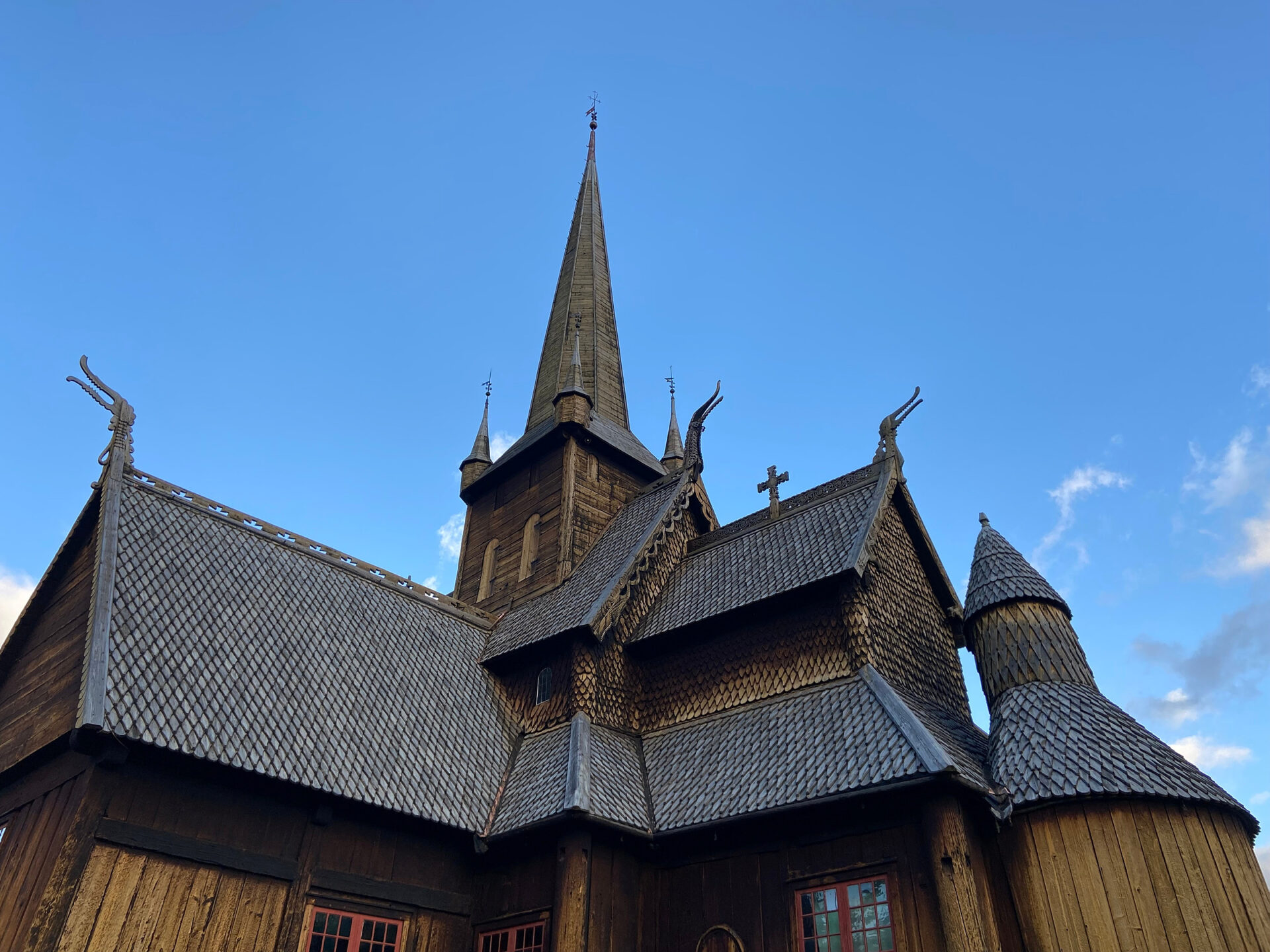 Stavkirke i tre med bratte spåntak og utsmykkede utskjæringer, toppet av et høyt spir mot en klarblå himmel.