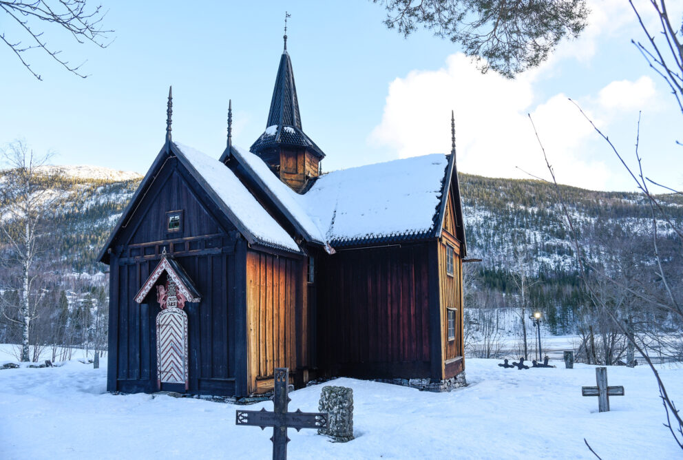 Milliontilskot til stavkyrkjene 7 En stavkyrkjene i tre med bratt tak står midt på en snødekt kirkegård i et fredfylt fjellandskap om vinteren.