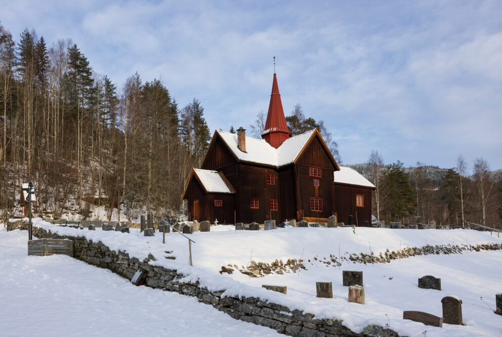 Milliontilskot til stavkyrkjene 6 En stavkyrkjene i tre med et rødt kirketårn står på en snødekt kirkegård omgitt av trær og en steinmur under en delvis overskyet himmel, og minner om kulturminner som nå er bevart ved hjelp av milliontilskuddet.