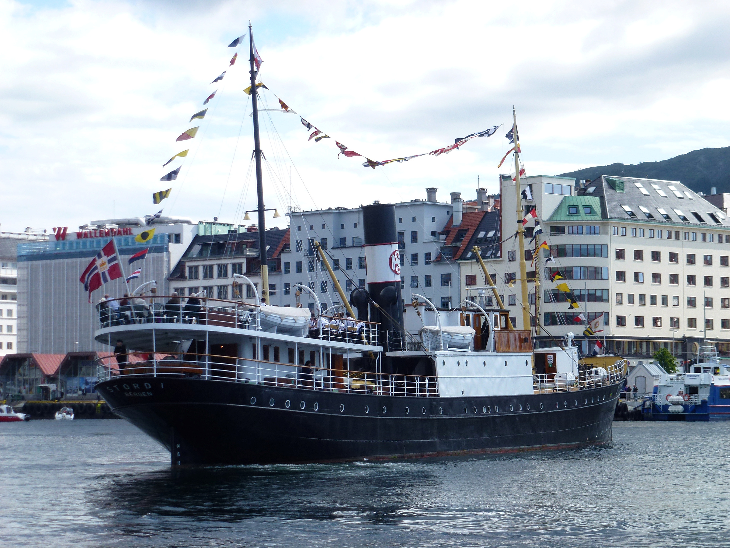 Et svart-hvitt dampskip dekorert med maritime flagg seiler nær en kai, med bygninger i flere etasjer og åser i bakgrunnen.