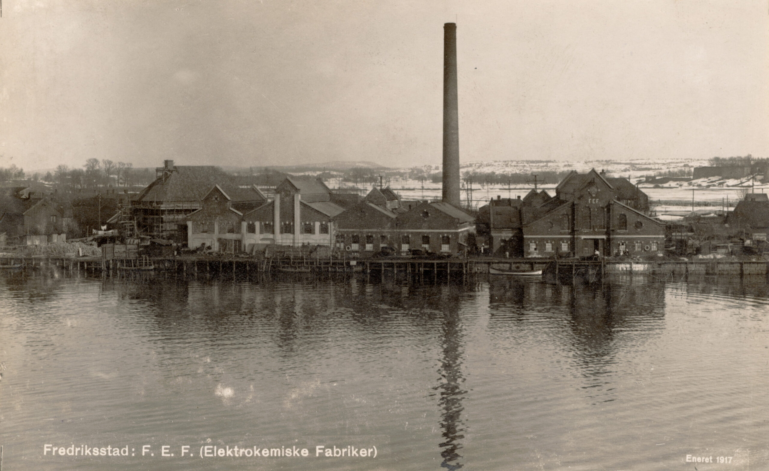 Svart-hvitt-foto av et industrikompleks med en høy skorstein ved en elvebredd, merket Fredrikstad: F. E. F. (Elektrokemiske Fabriker), tatt i 1917.