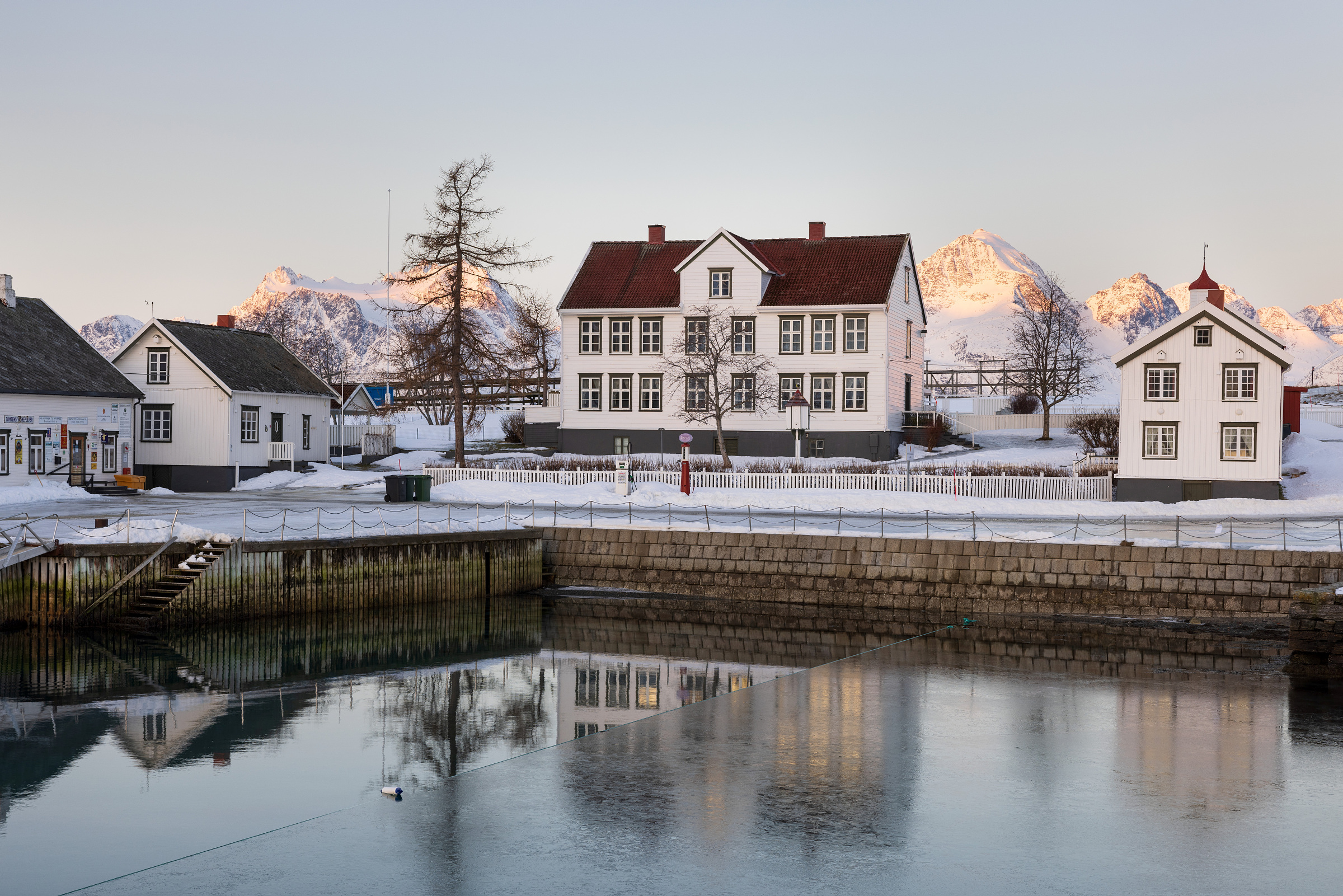 En scene ved vannkanten med hvite trehus, snødekt mark, løvløse trær og fjell i bakgrunnen - et idyllisk kulturmiljø som speiler seg i et rolig, delvis tilfrosset vann.