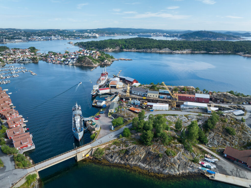 Flyfoto av et skipsverft med en ferge ved kai, omgitt av vann, hus, båter og skogkledde øyer under en blå himmel.