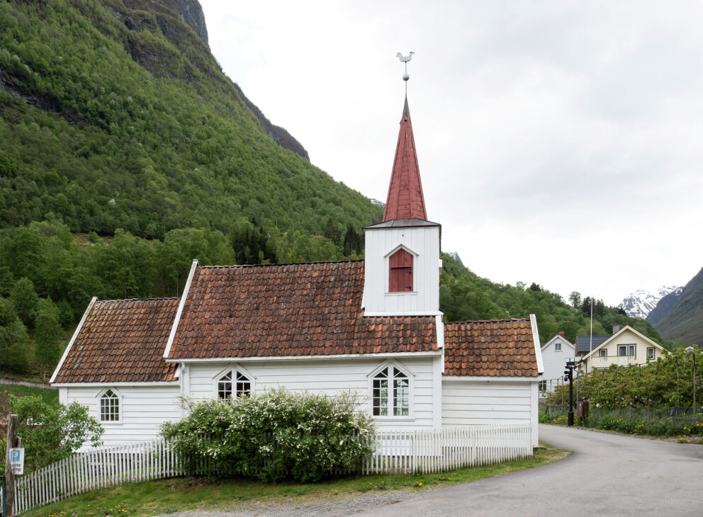 Milliontilskot til stavkyrkjene 10 En liten hvit trekirke med et rødt kirketårn og en værhane står ved en buet vei som minner om de tradisjonelle stavkyrkjene, omgitt av grønne åser og trær.